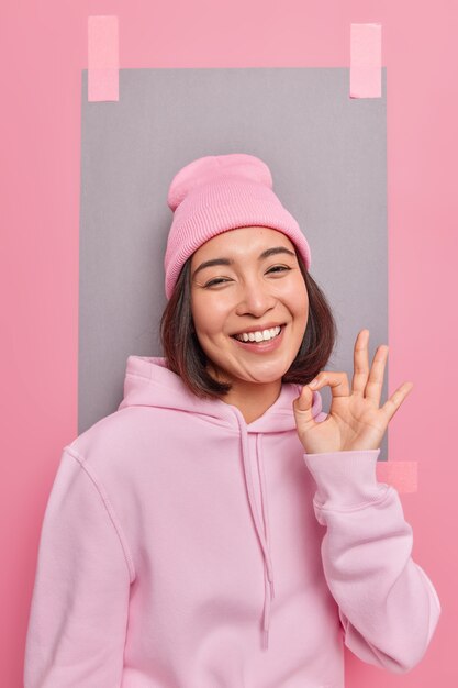 Cheerful carefree woman shows okay gesture in approval demonstrates recommendation sign agrees with you praises good work wears hoodie and hat poses indoor