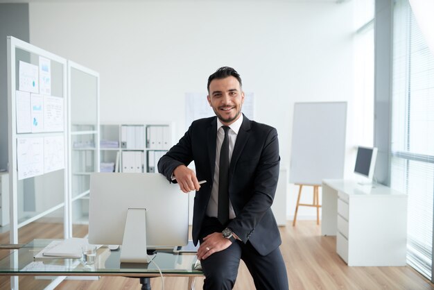 Cheerful Caucasian businessman sitting on desk in office, leaning on screen and smiling