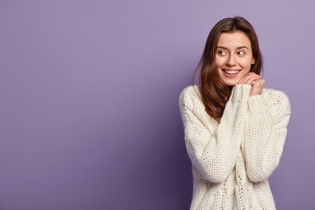 Cheerful Caucasian girl keeps hands together near face, looks positively aside, has no make up, healthy skin, wears white sweater, stands over purple wall with blank space for your promotion