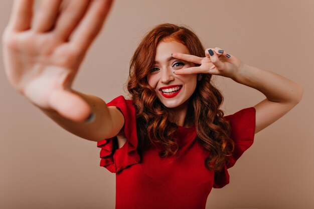 Cheerful caucasian girl in red dress dancing . Wonderful young woman with ginger hair posing with peace sign.