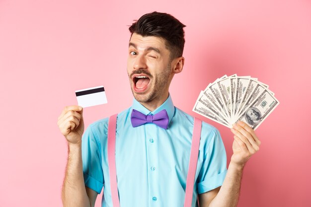 Free photo cheerful caucasian man in bowtie winking at you showing plastic credit card and money recommending s...