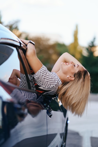 Cheerful Caucasian woman drives through the picturesque sunny city and waves her arms while stretching out of the car window on a beautiful day