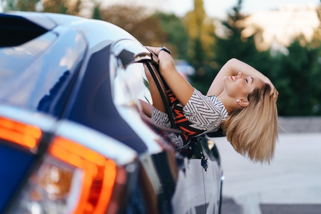 Cheerful Caucasian woman drives through the picturesque sunny city and waves her arms while stretching out of the car window on a beautiful day