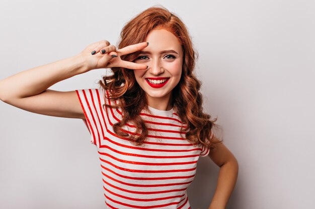 Cheerful caucasian woman in red t-shirt posing with peace sign. Indoor photo of ginger emotional girl laughing on white wall.
