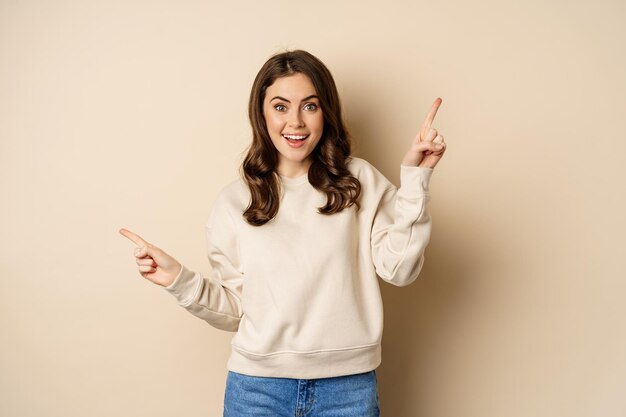 Cheerful caucasian woman showing directions, two ways, pointing sideways at variants, choices for customer, standing over beige background
