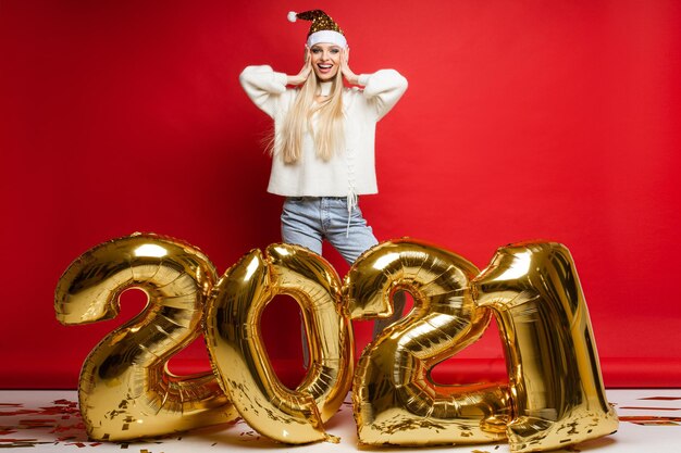 Cheerful caucasian young woman in white sweater, blue jeans and red christmas hat keeps her hands near the head and smiles, picture isolated on red background