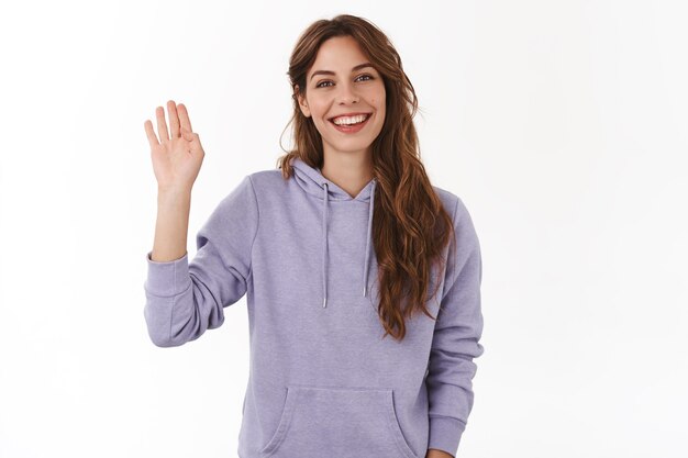 Cheerful charismatic attractive friendly woman waving raised hand hi greeting gesture smiling toothy perfect grin introduce herself meet new people welcoming guests, white wall
