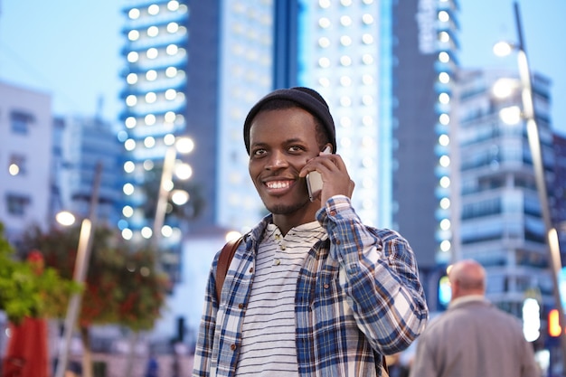 Cheerful charismatic young Afro American student in stylish wear smiling happily while having phone conversation with his old friend