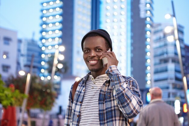 Cheerful charismatic young Afro American student in stylish wear smiling happily while having phone conversation with his old friend