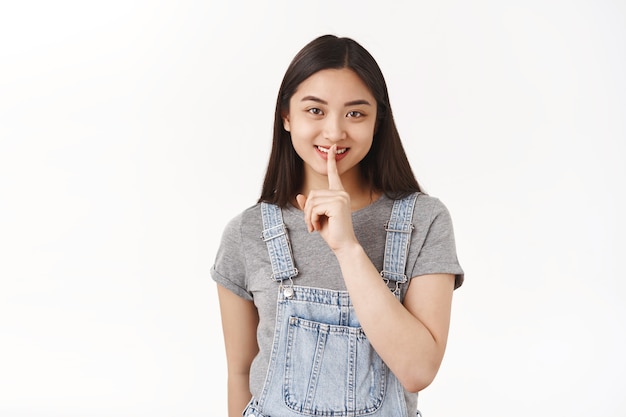 Cheerful cheeky cute asian brunette girl wear denim summer dungarees shush smiling cunning have plan share mystery ask keep quiet make surprise white wall