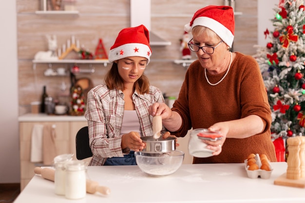 Cheerful child making traditional dough puttting flour in bowl using strainer cooking homemade gingerbread dessert with grandmother celebrating christmas season. Child enjoying winter holiday