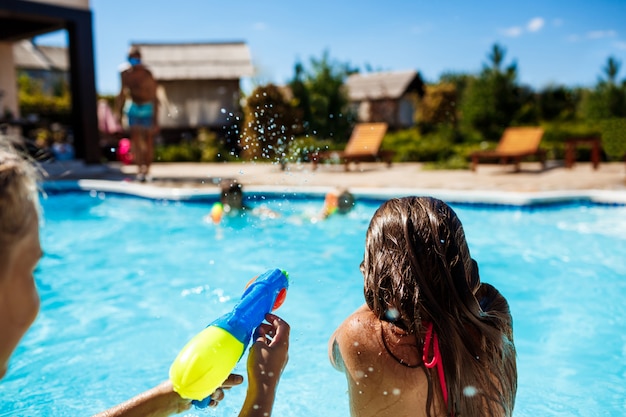 Cheerful children playing waterguns, rejoicing, jumping, swimming in pool.