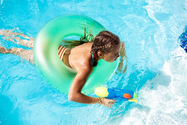 Cheerful children playing waterguns, rejoicing, jumping, swimming in pool.