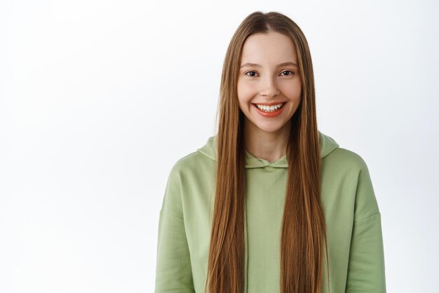 Cheerful and confident female model with long healthy hair natural makeup smiling and looking assertive at camera standing in green hoodie against white background