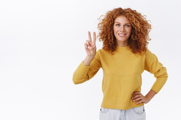 Cheerful, confident redhead curly woman in yellow sweater showing peace, goodwill sign and smiling determined, hold arm on hip, aim to win, feeling self-assured and assertive, white wall