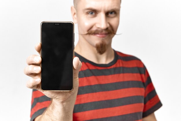 陽気な自信を持って若いビーズの男が幸せそうに笑って、完璧なデザインとコピースペースのディスプレイを備えたモダンな真新しい黒の携帯電話を示しています。電子機器で手に選択的な焦点