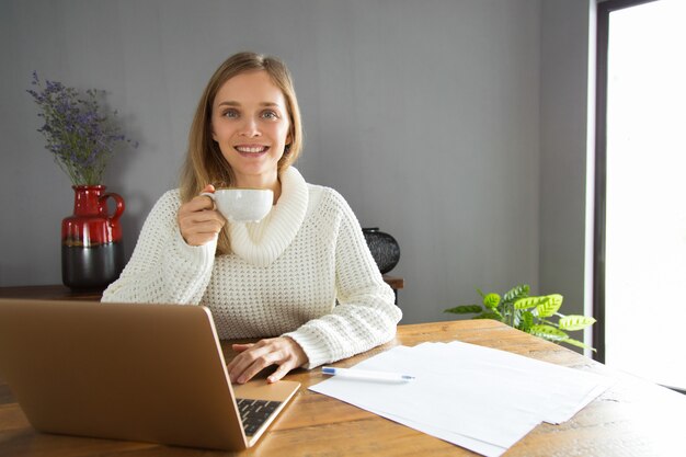 Веселая уверенно молодая женщина, используя ноутбук и пить кофе