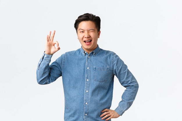 Cheerful confindent asian man have all under control, wink encouraging and show okay gesture, approve plan, congratulating person, saying well done, rate excellent job, standing white background.