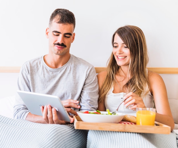 Cheerful couple eating salad and watching film on tablet