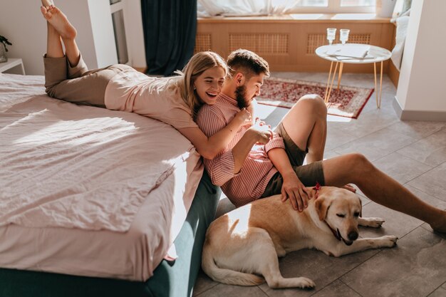 Cheerful couple is having fun in bedroom and playing with dog lying on floor. Girl laughs and hugs boyfriend.