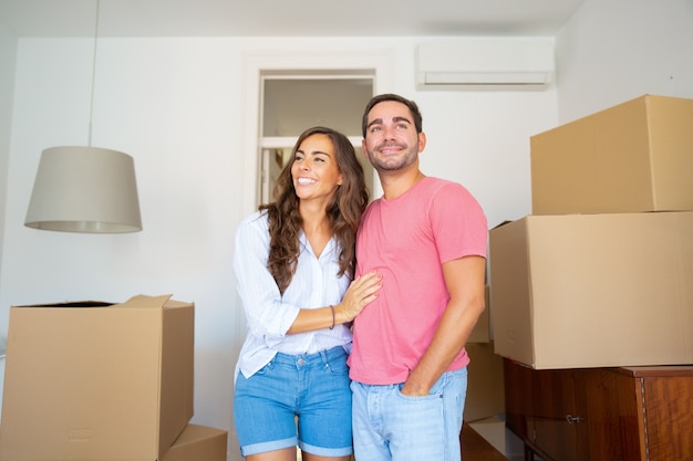 Cheerful couple looking over their new apartment, walking among carton boxes and hugging