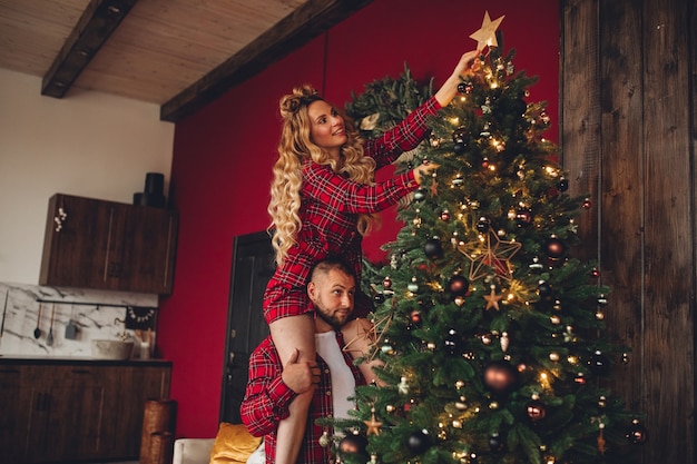 同じパジャマで恋をしている陽気なカップルが家で一緒にクリスマスツリーを飾ります