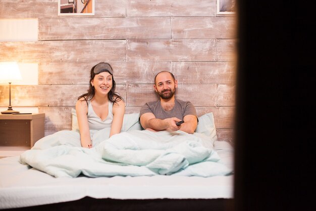 Cheerful couple in pajamas watching a comedy movie on big tv.