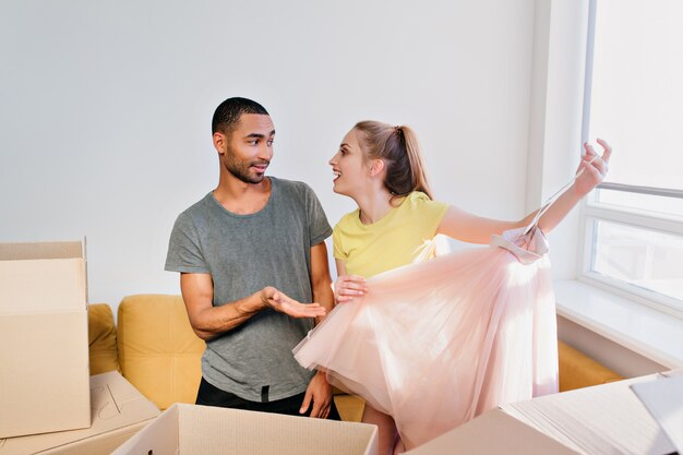 Cheerful couple unpacking boxes, family moved in new house, bought apartment. Young woman unboxing clothes, holding pink skirt. Wife and husband in room, wearing T-shirt, yellow top, shorts.