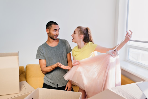 Веселая парочка распаковывает коробки, семья переехала в новый дом, купила квартиру. Молодая женщина распаковывает одежду, держа розовую юбку. Жена и муж в комнате, в футболке, желтом топе, шортах.