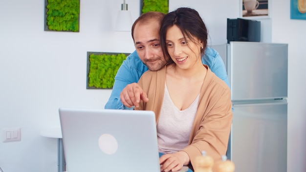 Cheerful couple using laptop in kitchen reading online recipe for breakfast. Husband and wife cooking recipe food. Happy healthy together lifestyle. Family searching for online meal. Health fresh sala