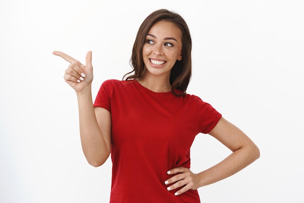 Веселая любопытная женщина в красной футболке, стоящая уверенно, держит руку на бедре, поворачивает голову влево, указывая пальцем в сторону, проверяя потрясающее место, стоя на белом фоне, с энтузиазмом представляет промо