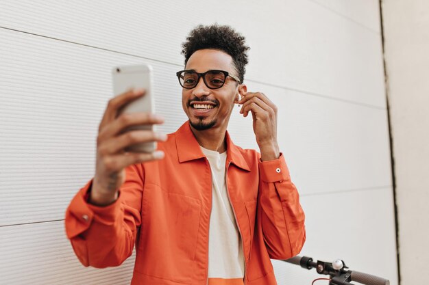 Cheerful curly darkskinned man in orange jacket and white tshirt smiles takes selfie and puts on wireless headphones outside