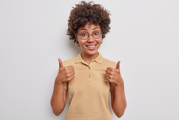 陽気な巻き毛の女の子は親指を立て続けるように表現し、正のフィードバックは承認し、計画の賞賛をサポートします店の販売は白い背景の上に分離されたカジュアルなTシャツを着て良い結果を祝います