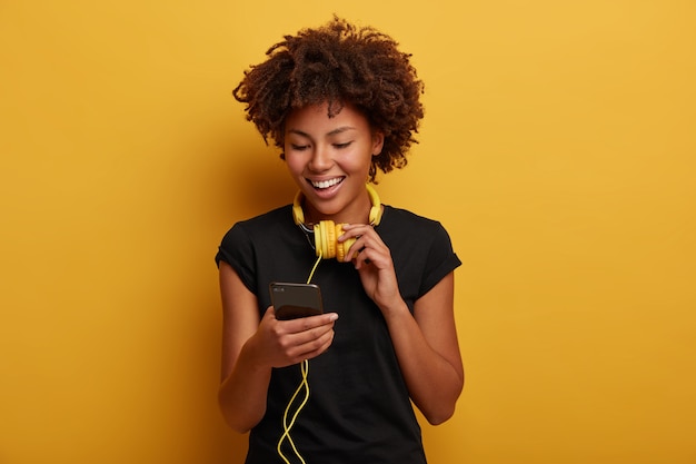 Cheerful curly girl picks up song in playlist, wears yellow headset around neck