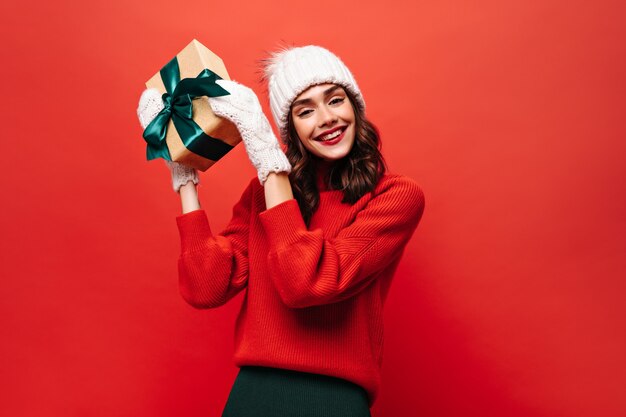 Cheerful curly girl in white warm hat, mittens and red sweater shakes gift box and smiles on red wall