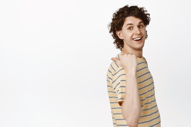 Cheerful curly guy pointing finger left smiling at camera showing logo or sale banner making announcement white background