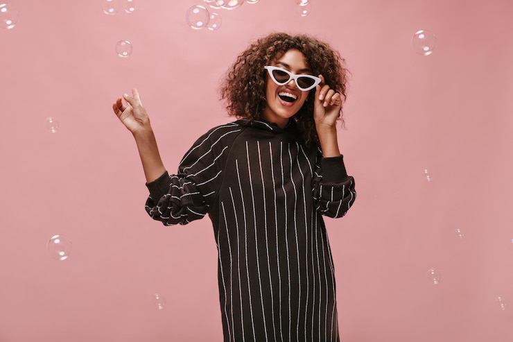 Free Photo cheerful curly haired girl in striped black dress with long sleeve and stylish light sunglasses laughing and posing with bubble on pink backdrop