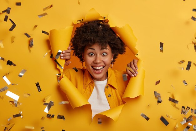 Cheerful curly haired lady with toothy smile, wears fashionable clothes and silver earrings, looks through paper hole with bright sparkles