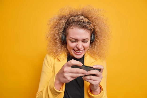 Бесплатное фото Веселая кудрявая женщина, увлекающаяся видеоиграми на мобильном телефоне, использует беспроводные наушники, сосредоточенные на дисплее, наслаждается играми, одетая формально изолированно над ярко-желтой стеной