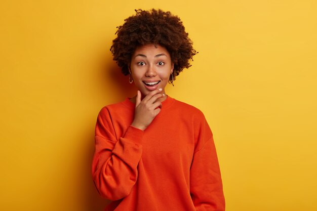 Cheerful curly haired woman holds chin, expresses bright emotions