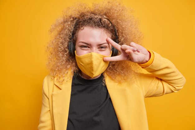 Cheerful curly haired woman makes peace gesture over eye has fun listens music via headphones wears protective mask during pandemic poses against vivid yellow wall. Body language concept