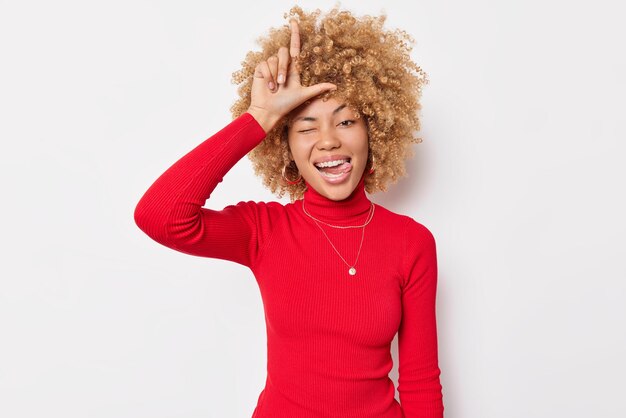 Cheerful curly haired woman mocks friend being last makes loser gesture over forehead sticks out tongue wears casual red turtleneck isolated over white background. Outsiders should stay at home