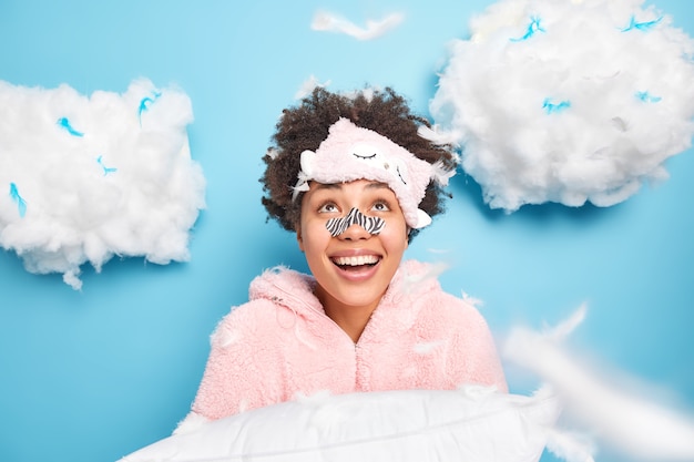 Cheerful curly haired young woman wears applicator mask on nose dressed in nightwear looks gladfully above smiles broadly holds pillow prepares for sleep and having rest isolated over blue wall