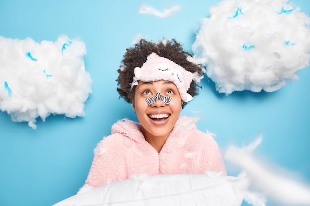 쾌활한 곱슬 머리 젊은 여자는 잠옷을 입은 코에 어플리케이터 마스크를 착용하고 미소 위에 기꺼이 보이는 베개는 수면을 준비하고 나머지는 파란색 벽 위에 절연되어 있습니다.