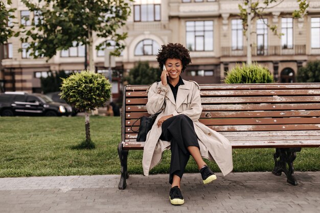 Cheerful curly lady in black pants and beige trench coat sits on wooden bench outdoors