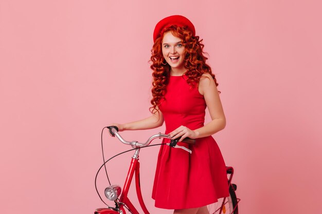 빨간 드레스와 세련된 모자를 쓴 쾌활한 곱슬머리 아가씨가 웃고 있다 자전거의 운전대를 들고 있는 소녀의 초상화