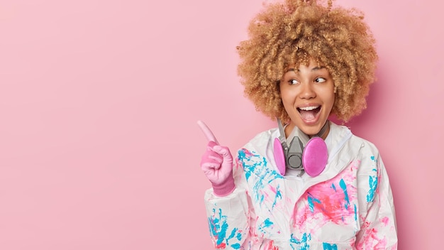 Cheerful curly woman wears hazmat suit gloves and respirator around neck points away on blank space against pink background demonstrates place for your advertisement Chemical protection concept