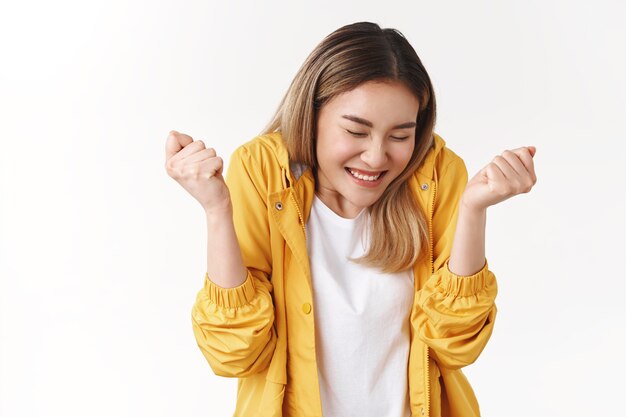 陽気なかわいいアジアのブロンドの女の子は大喜び幸せを感じます甘い成功ダンス目を閉じて笑顔を広く握り締める手を勝利を祝う優れたニュース完璧なスコアは目標を達成します