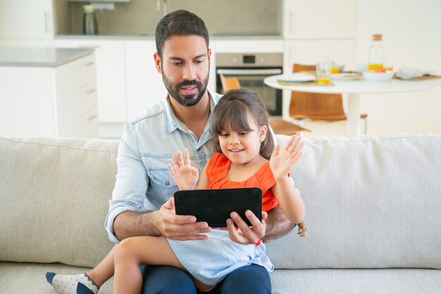 Cheerful cute girl and her dad using online app or watching footage on tablet together.