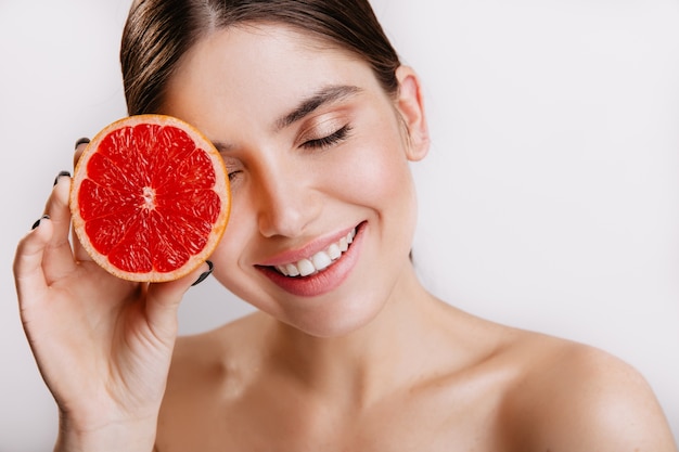 白い壁に赤い健康的な柑橘系の果物でポーズをとって、笑顔の陽気なかわいい女の子。